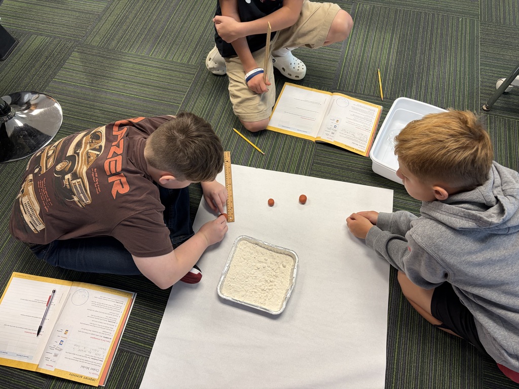 An overhead view of three students sitting on the floor measuring clay spheres with a wooden ruler next to a flour pan science setup.