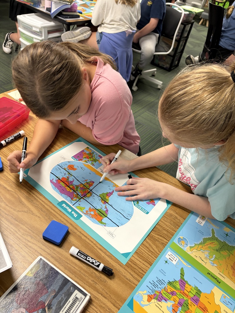 Two girls sitting side by side at a wooden desk drawing lines across a laminated world map with black dry-erase markers.