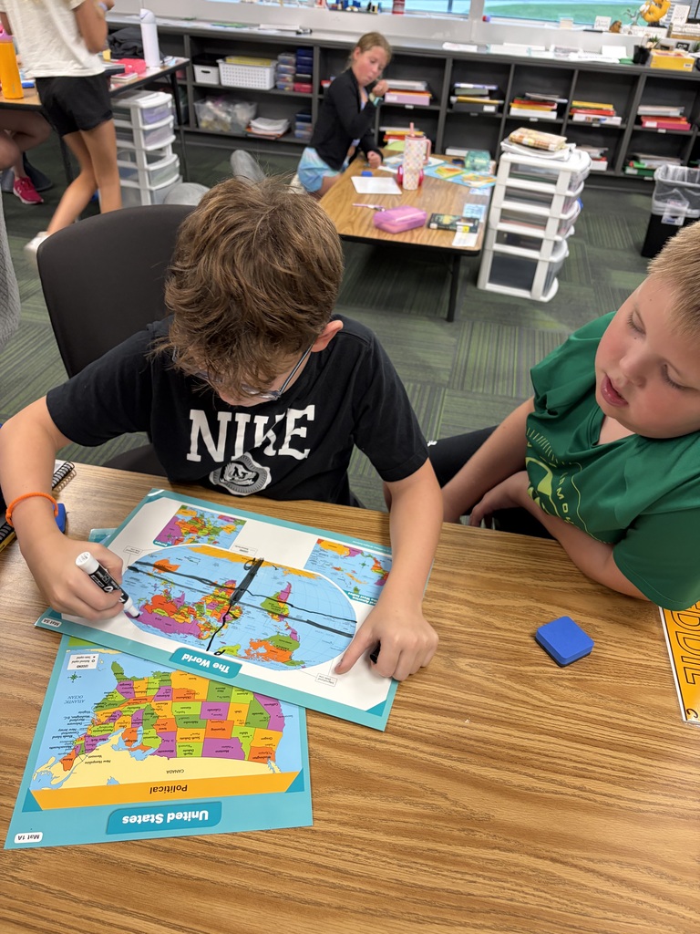 Two students leaning over a desk while tracing longitude and latitude lines onto a world map laminate.