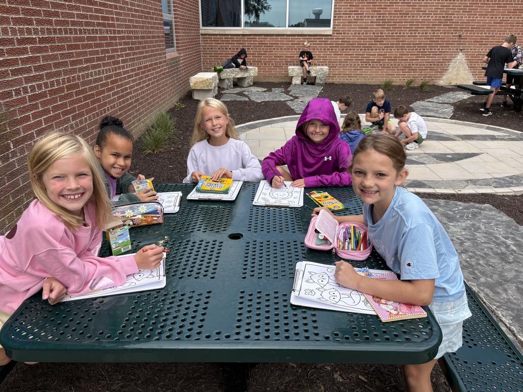 Five young students sitting around a green outdoor picnic table outside a brick school building, smiling while coloring on clipboards with markers and crayons.