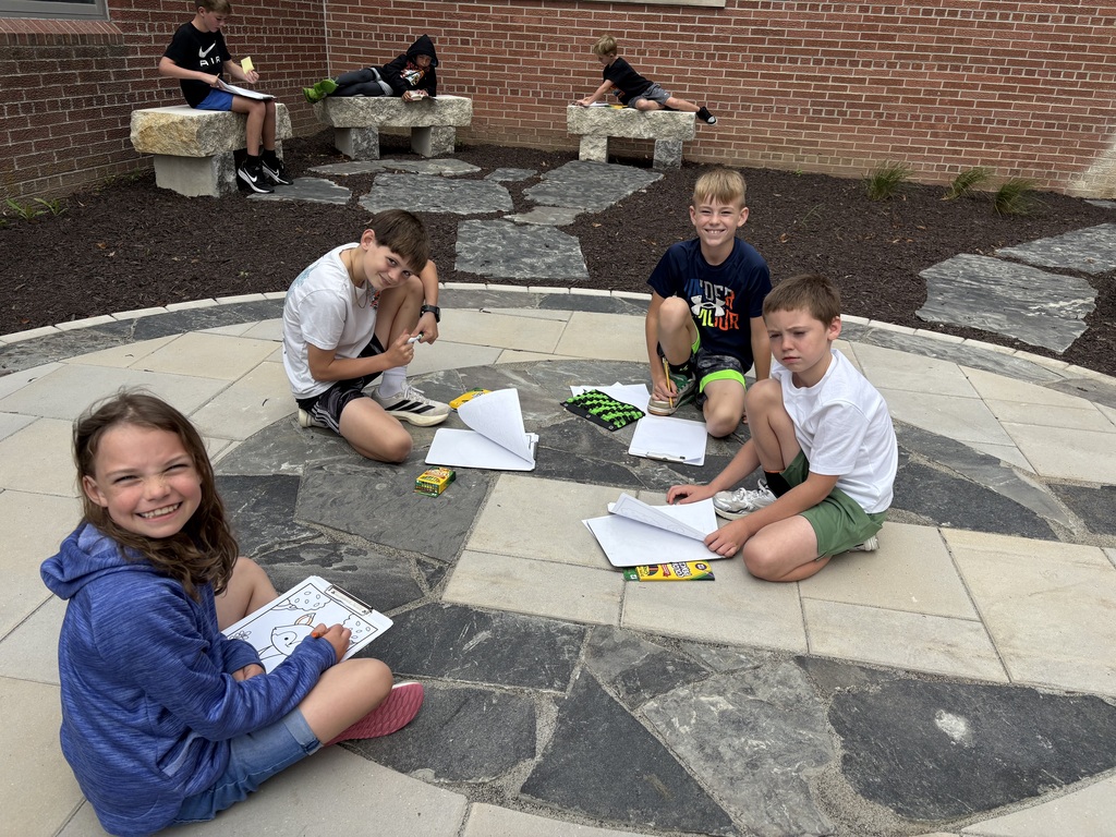 Four children sitting on a stone patio outdoor courtyard, working on coloring pages and schoolwork spread out around them.