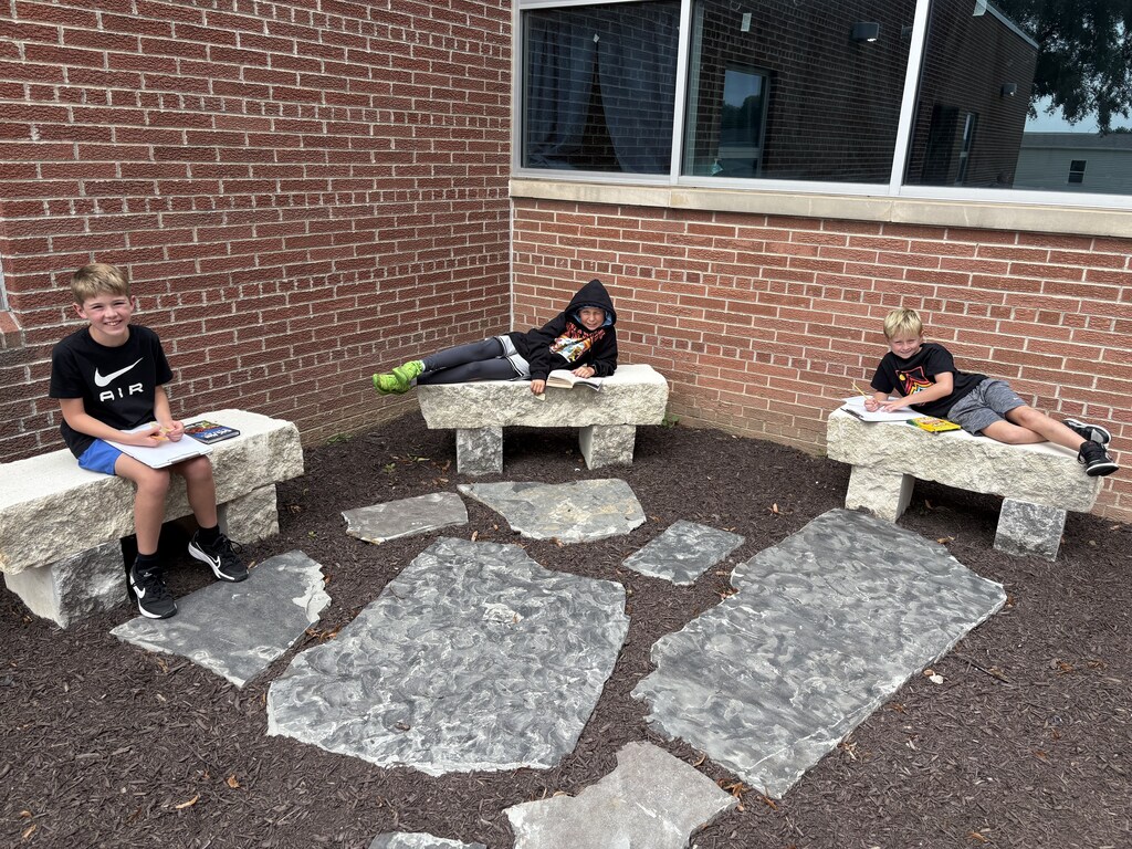 Three students sitting and lounging on stone outdoor benches near a brick wall, reading books and writing on clipboards.