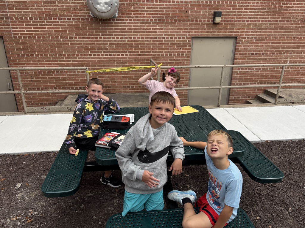 Four young students gathering around a green picnic table outside a school building, posing playfully for the camera.