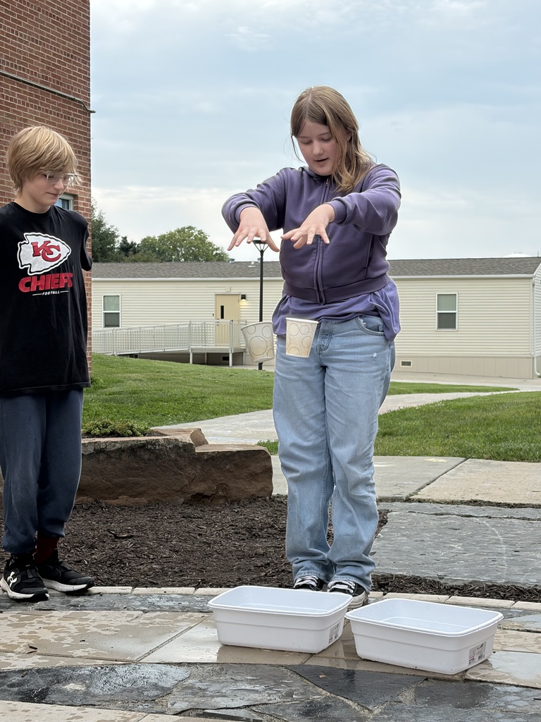 A girl dropping paper cups into plastic water bins outside while another student watches.
