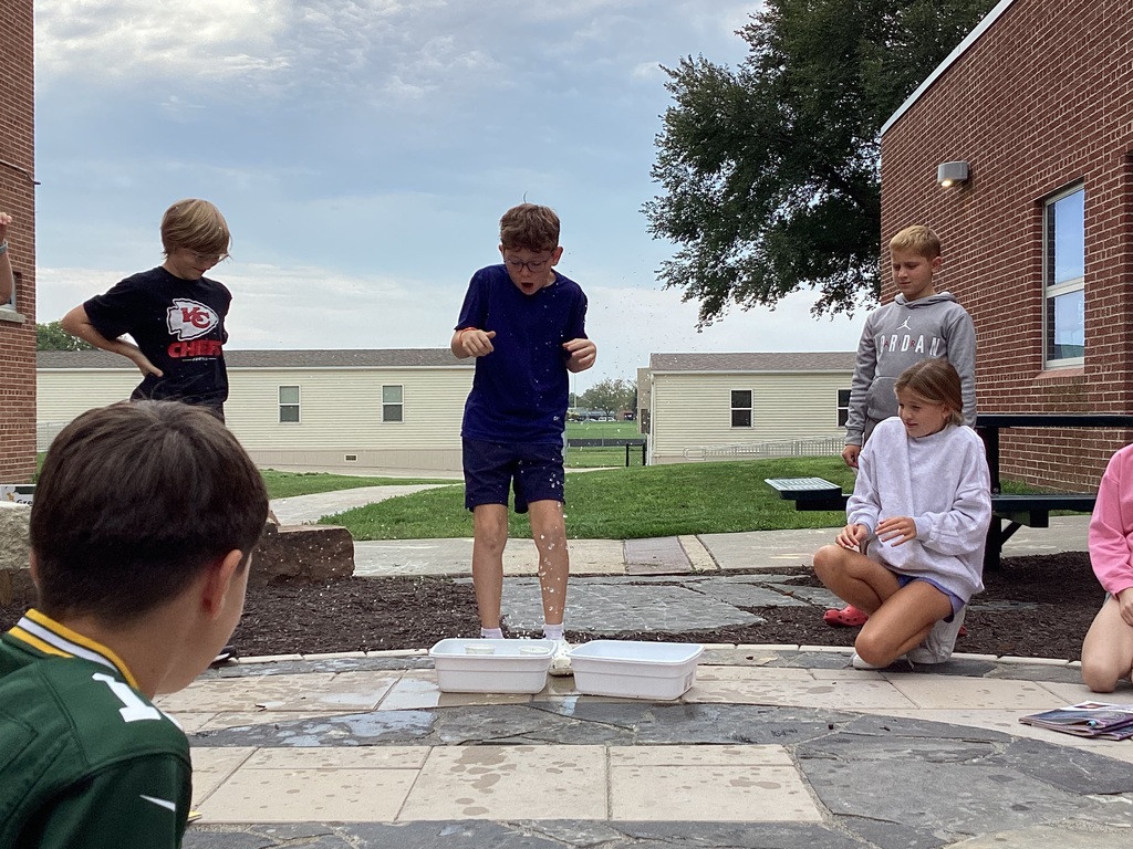 A group of elementary students gathered outdoors watching a boy conduct a science experiment with plastic tubs and water.