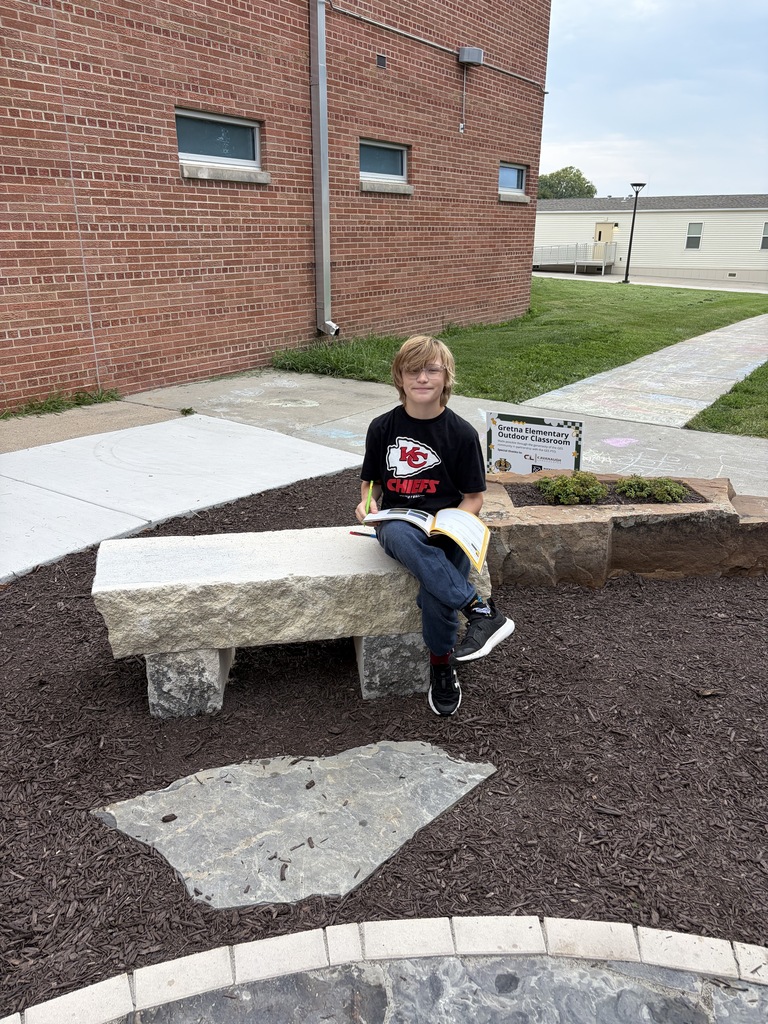 A young boy sitting on a stone bench outdoors, smiling while holding an open workbook and pencil.