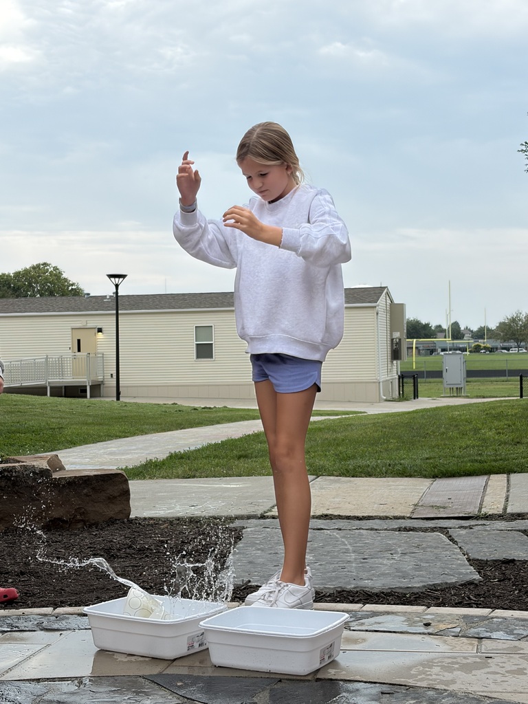 A young girl in a white sweatshirt dropping paper cups into a plastic tub of water outdoors.
