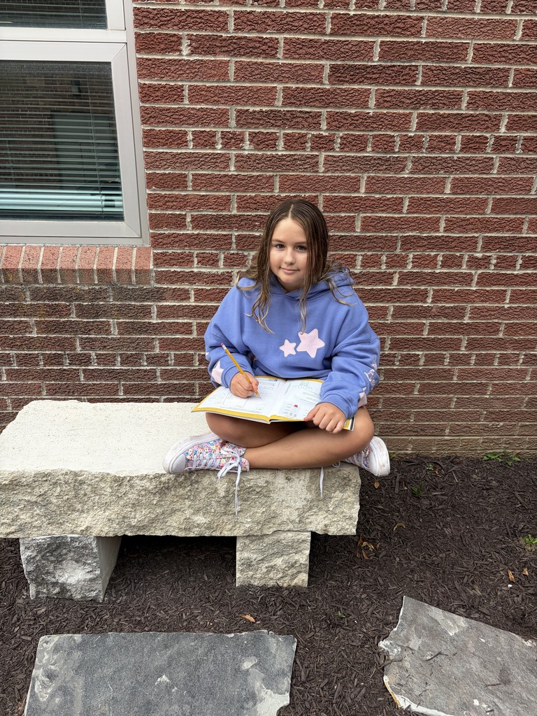 A young girl wearing a blue hoodie sitting cross-legged on a stone outdoor bench while writing in a notebook.