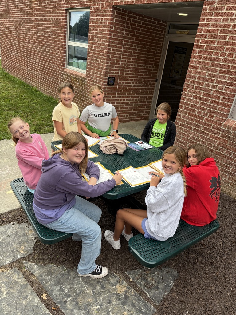 Seven young girls seated around a green outdoor picnic table working together on schoolwork.