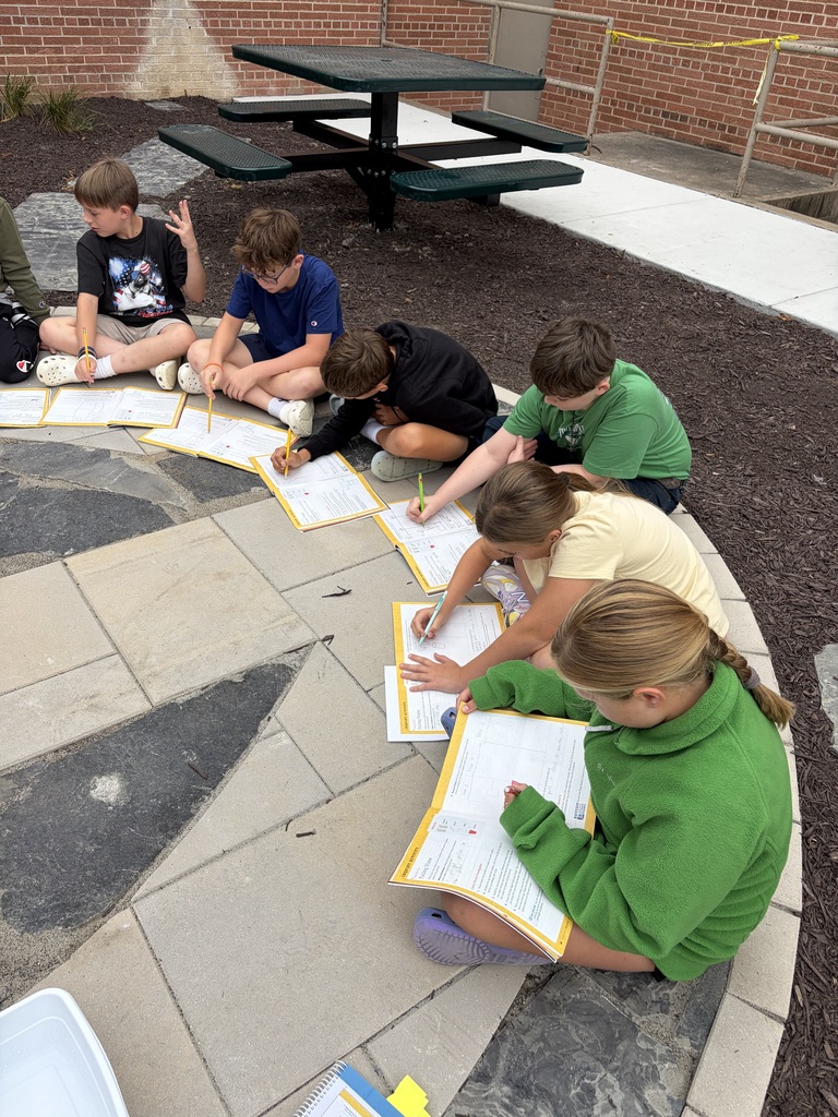 A semi-circle of elementary students sitting on a stone patio outside, writing in yellow workbooks with pencils.