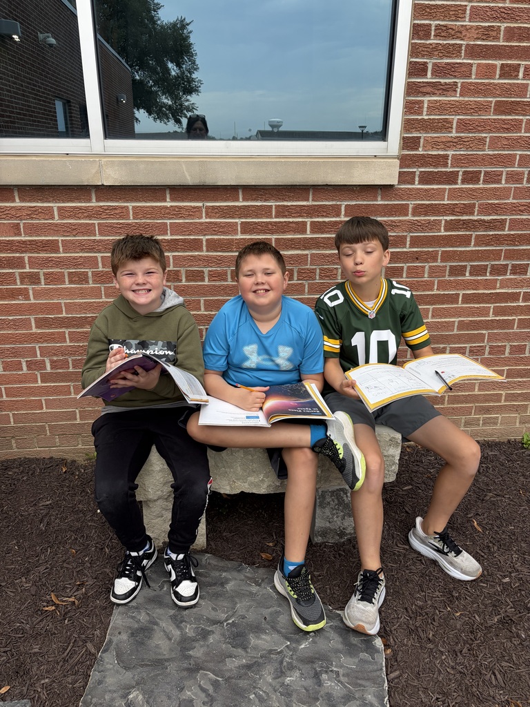 Three young boys sitting together on a stone bench outside a brick school building, holding open workbooks and smiling for the camera.