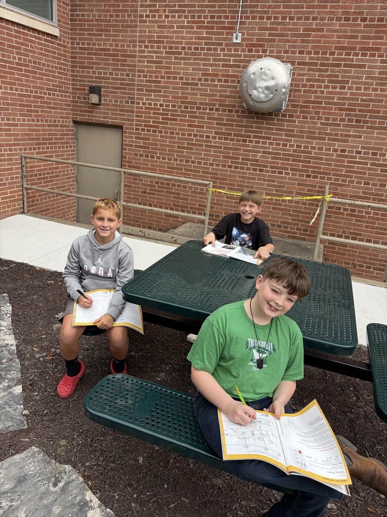 Three young boys sitting at a green outdoor picnic table writing in their yellow workbooks.