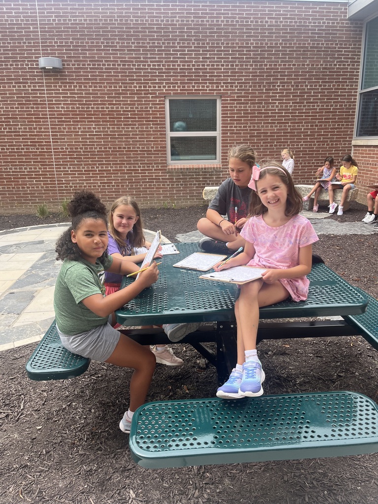 Four young girls seated around a green picnic table, smiling and writing on clipboards during outdoor class time.