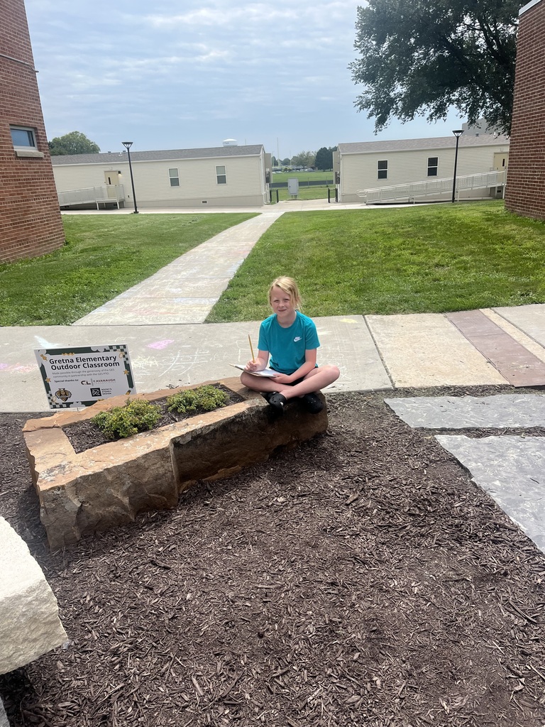 A student sitting cross-legged on a large stone planter in an outdoor classroom area while writing on a clipboard.