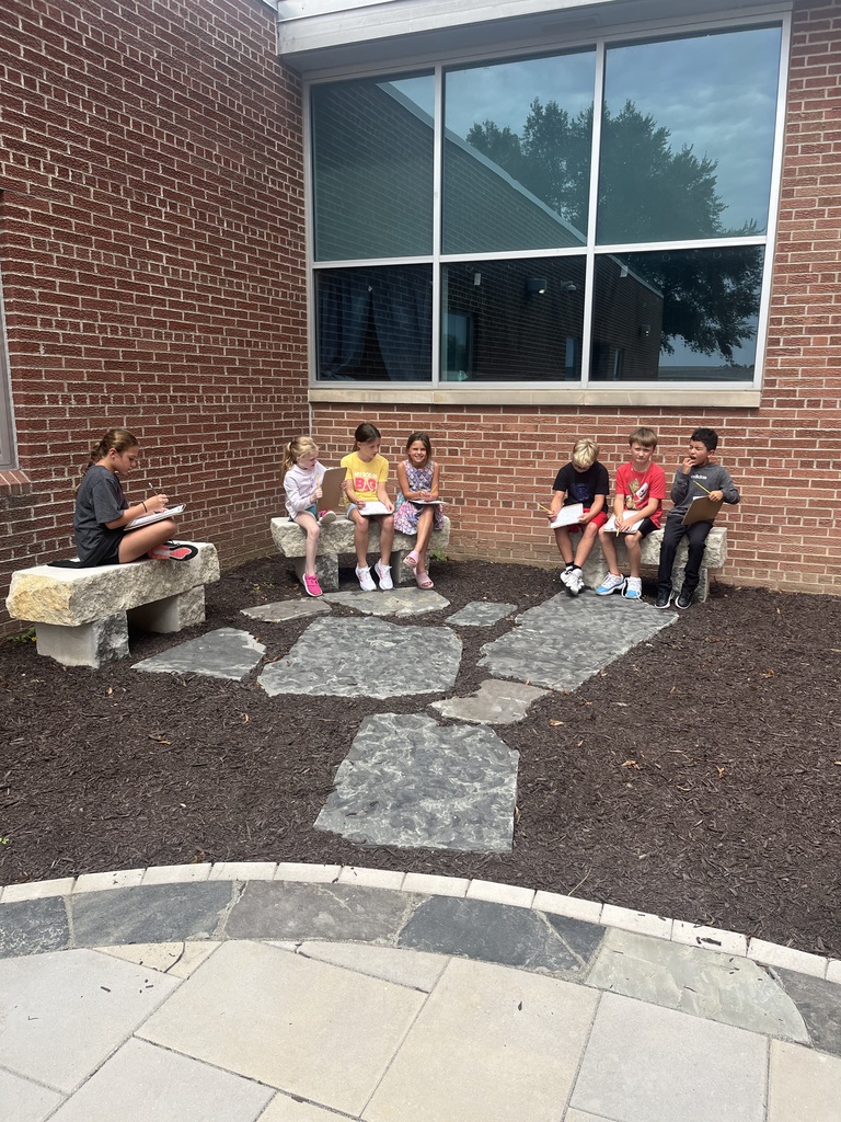 Seven students sitting along outdoor stone benches against a brick wall, writing on clipboards.