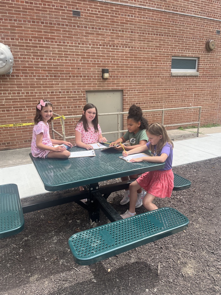 Four young students sitting around a green outdoor picnic table outside a brick building, filling out papers on clipboards.