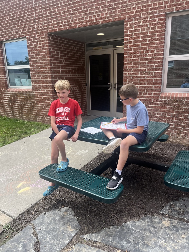 Two boys sitting at a green picnic table outside a school entrance, working together on clipboards.
