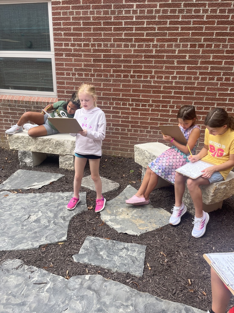 Four students in an outdoor courtyard area, with one girl standing and reading from a clipboard while others write on stone benches nearby.