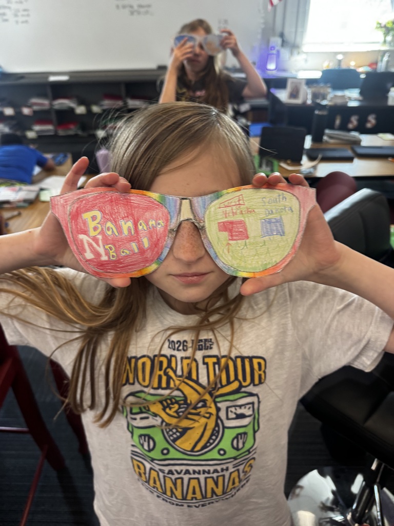 A girl holding up colorful paper cutout sunglasses with text reading "Banana Ball" on one side and state maps labeled "West Nebraska" and "South Dakota" on the other side.