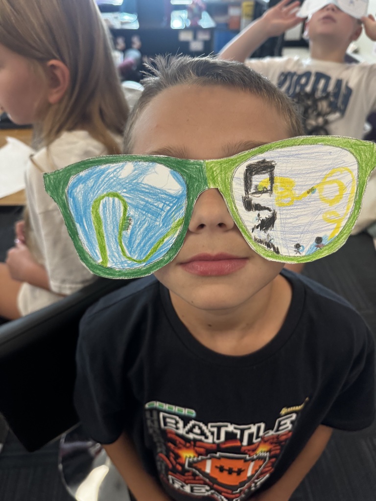 A student holding up bright green paper cutout sunglasses featuring hand-drawn illustrations of a roller coaster and amusement park rides.