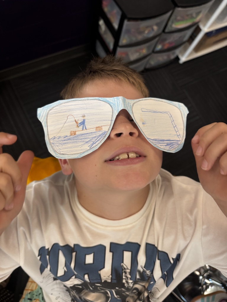 A boy looking up into the camera while holding up paper cutout sunglasses that feature hand-drawn scenes of fishing from a dock and a crane.