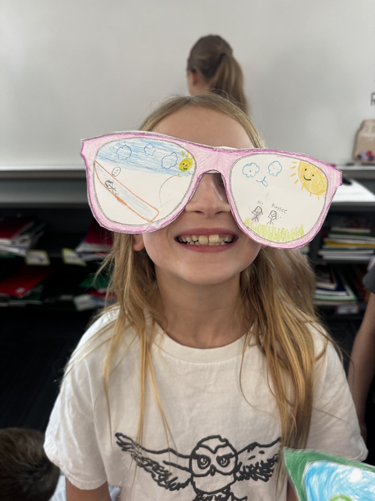 A smiling girl holding pink paper cutout sunglasses featuring drawings of a water slide, sunshine, and stick figures labeled "Me" and "Haylee."