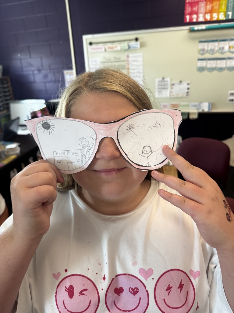 A student in a smiley-face shirt holding pink paper cutout sunglasses decorated with detailed pencil drawings of a building labeled "Five Below" and figures under the sun.