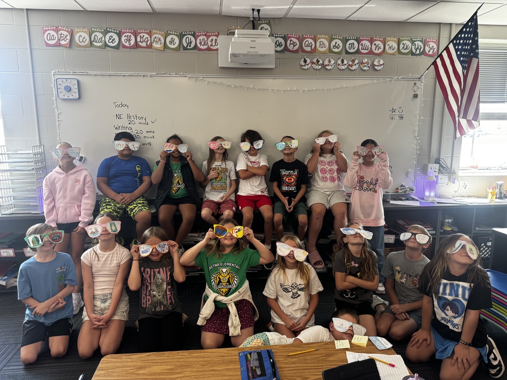 A full classroom group photo of students seated together, each holding up or wearing custom paper cutout sunglasses in front of a whiteboard.
