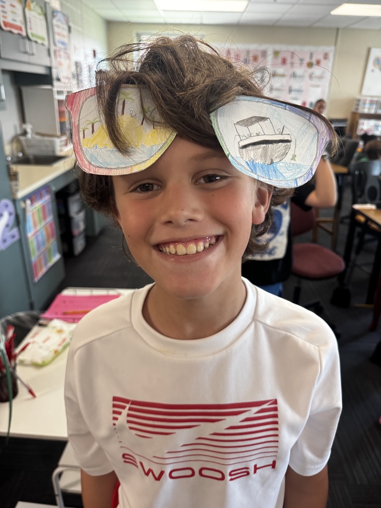 A smiling boy in a white Swoosh shirt wearing large paper cutout sunglasses decorated with drawings of a tropical island and a boat on the ocean.