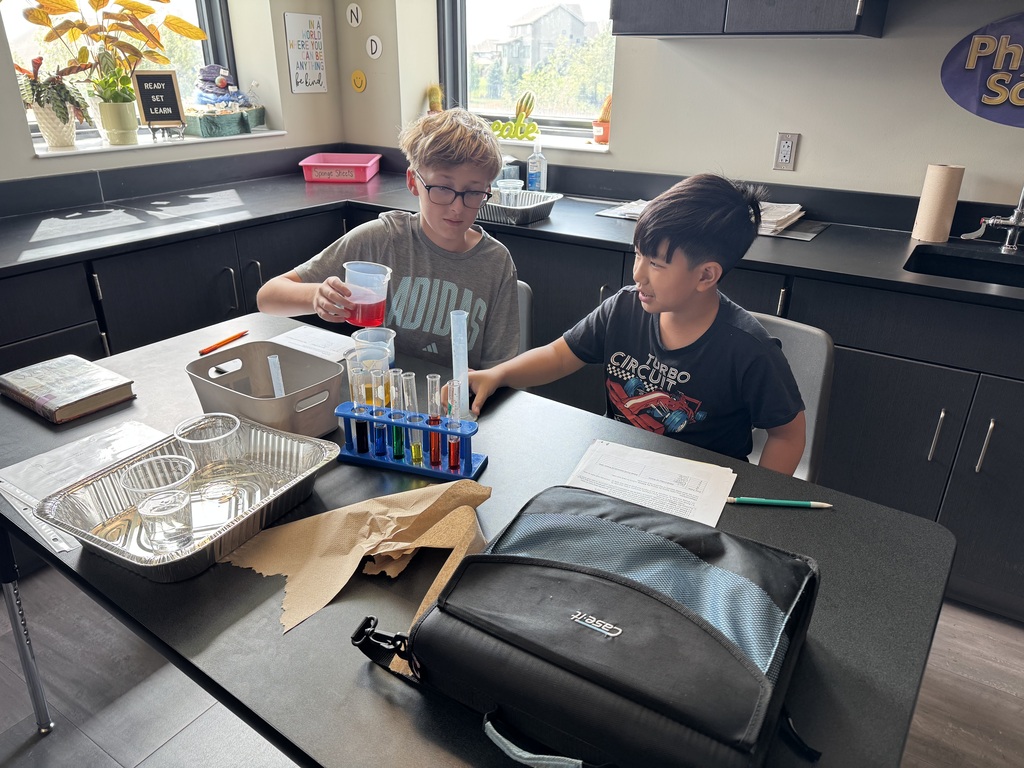Photo of two male students sitting at a table. They have several graduated cylinders in front of them filled with different colored liquids. The boy on the left is holding a cup of red liquid.
