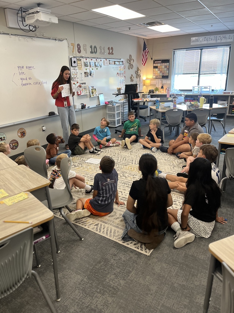 teacher explaining an assignment to students seated on the carpet