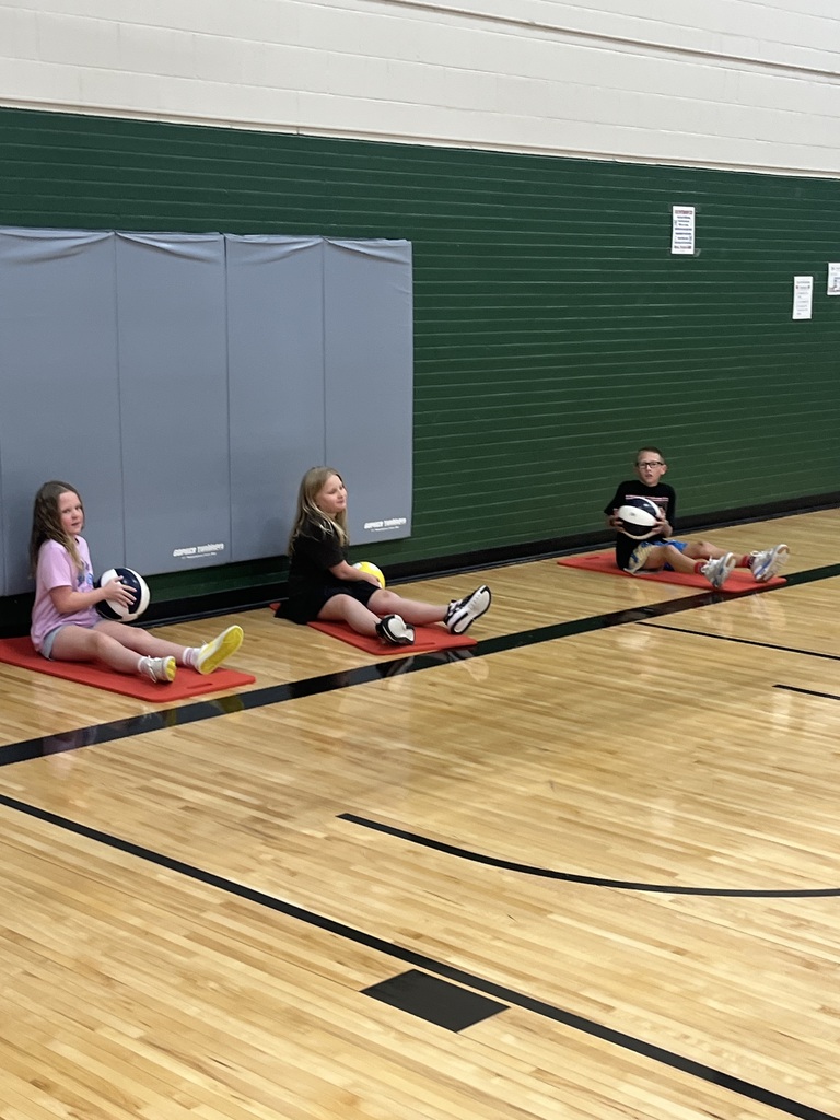 Three kids sitting on red mats doing Russian twists with basketballs along a gymnasium wall.