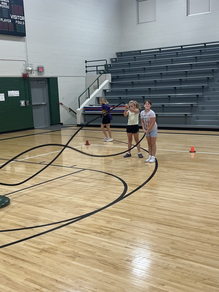 Two girls standing on a basketball court holding a battle rope while a third girl runs in the background.