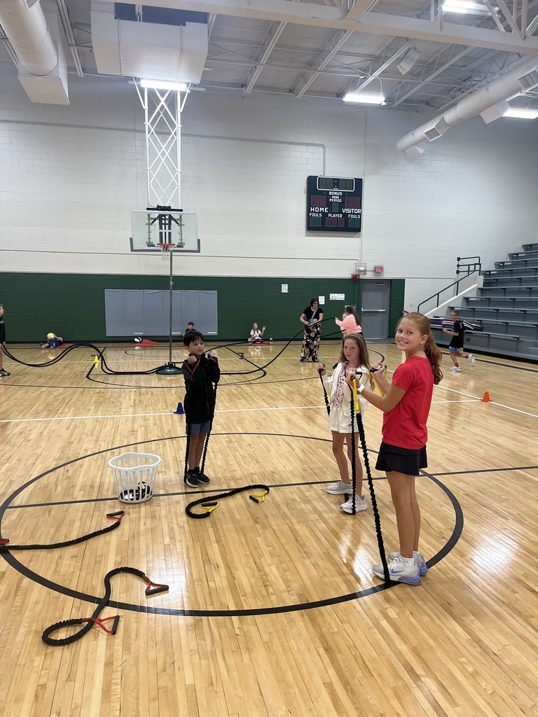 Three kids holding resistance bands attached to a basketball hoop pole during gym class.