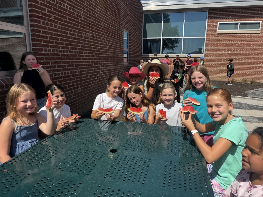 A group of young girls outdoors sitting at a green metal picnic table, smiling and holding up slices of watermelon.