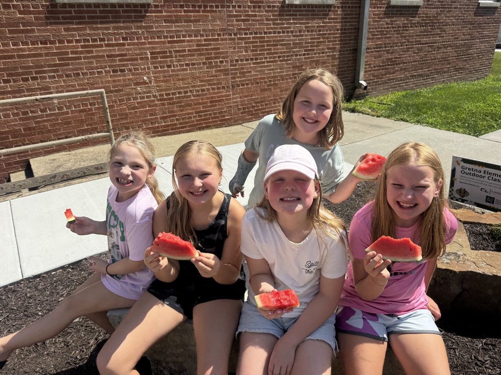 Five young girls sitting together outdoors on stone blocks, holding watermelon slices and smiling at the camera.