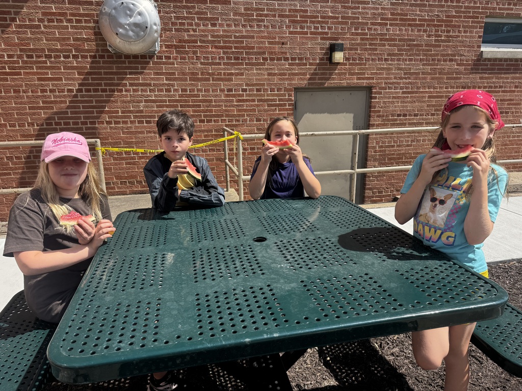 Four children sitting at a green metal outdoor picnic table, eating watermelon slices in front of a brick wall.