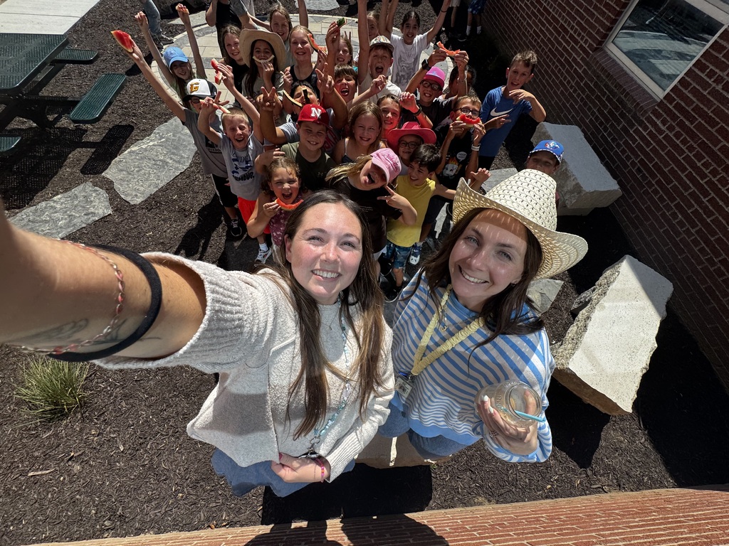 A high-angle selfie of two women smiling, with a crowd of cheerful students waving and holding up watermelon slices behind them in an outdoor courtyard.