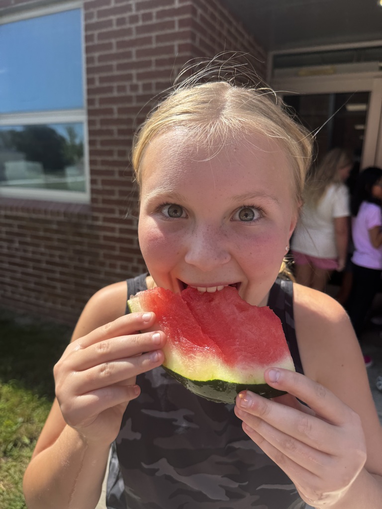 A close-up of a young girl with blonde hair taking a bite out of a large watermelon slice.