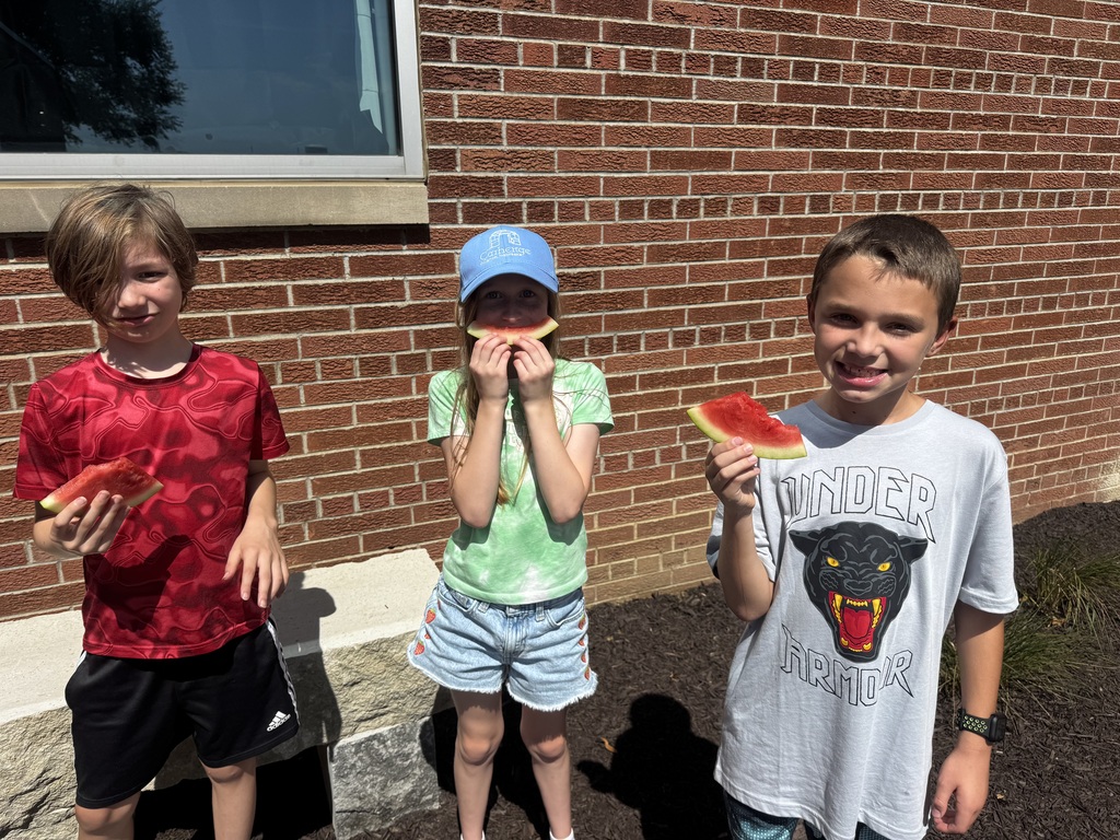 Three children standing side by side outside against a brick wall, each holding a slice of watermelon.
