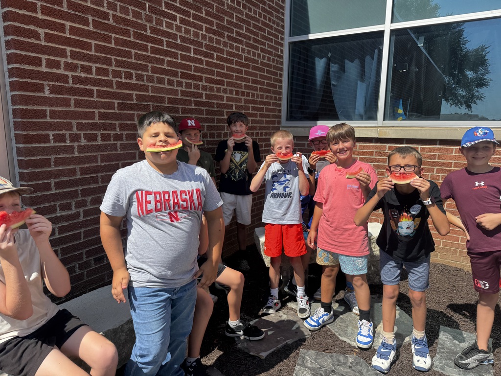 A group of young boys outdoors holding up watermelon slices near their faces, posing in front of a brick building.