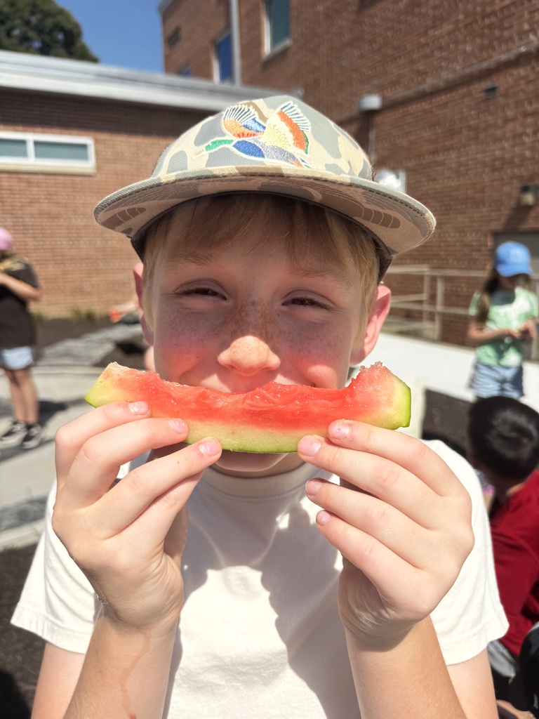 A close-up shot of a boy wearing a camo hat holding a watermelon slice up like a smile over his mouth.