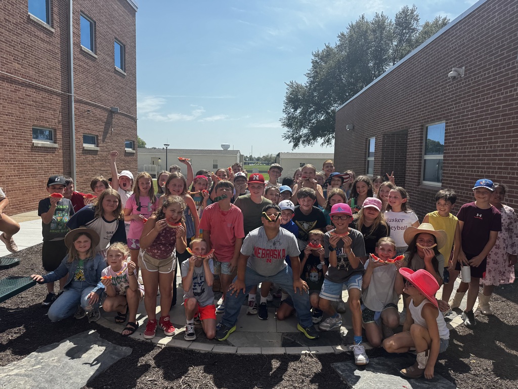 A large group of children outdoors posing together in a courtyard area between brick school buildings, holding up watermelon slices.