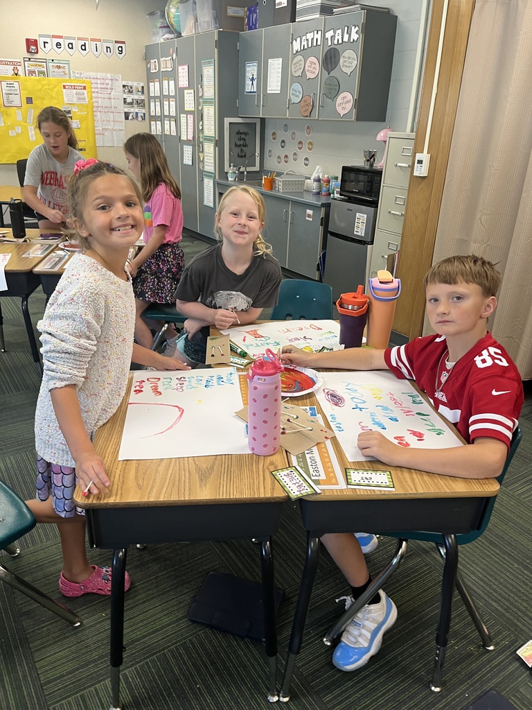 Four young students sitting and standing around classroom desks with colorful paint and artwork laid out in front of them.