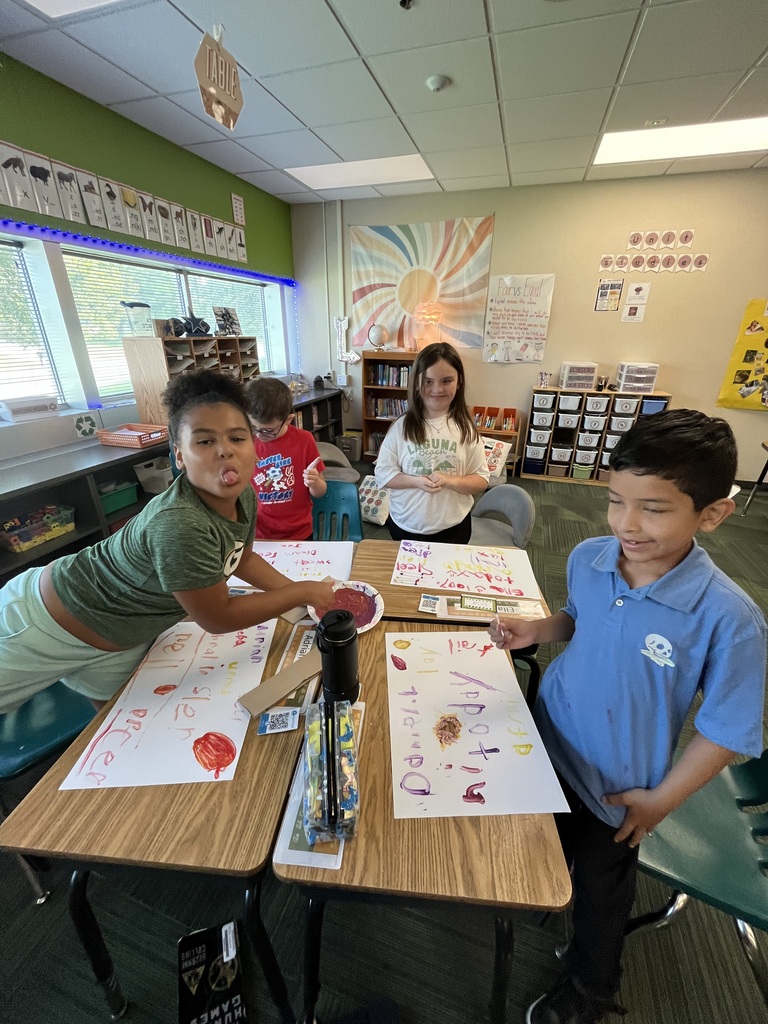 Students working on painting art projects at their classroom desks, with one student playfully sticking out their tongue at the camera.