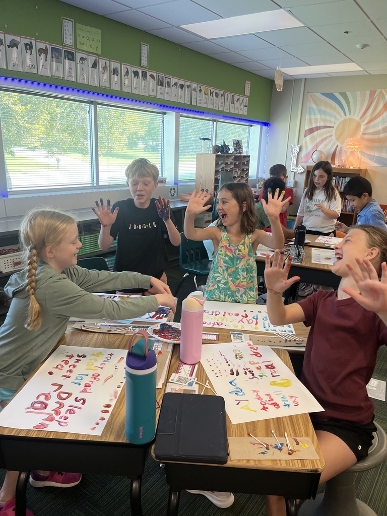 Elementary students sitting around connected classroom desks, excitedly raising paint-covered hands above their colorful art projects.