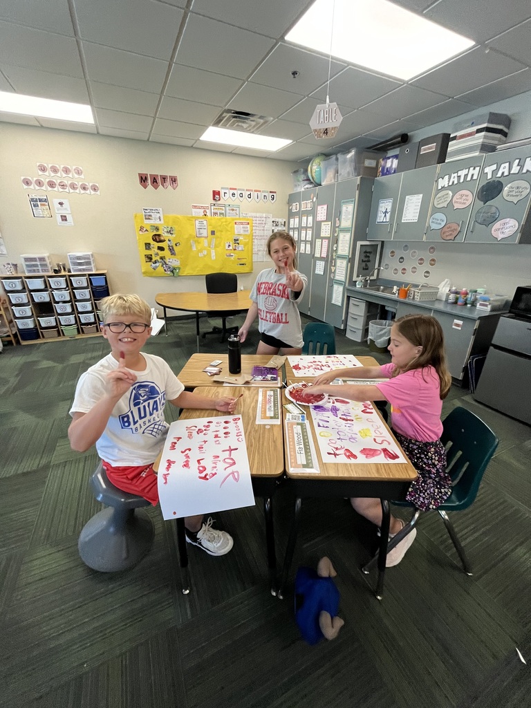 Three students sitting at desks in a classroom, with two holding up paint-covered fingers and painting on paper sheets.