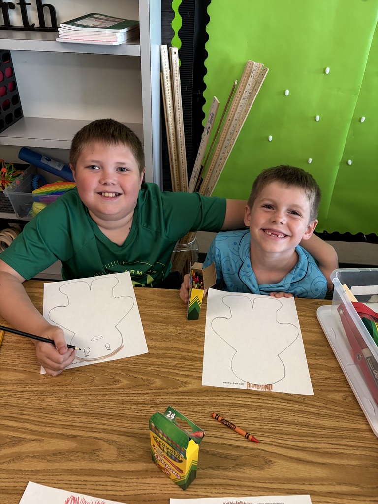 Two smiling boys sitting side-by-side at a wooden table, coloring in their gingerbread person outlines with crayons.