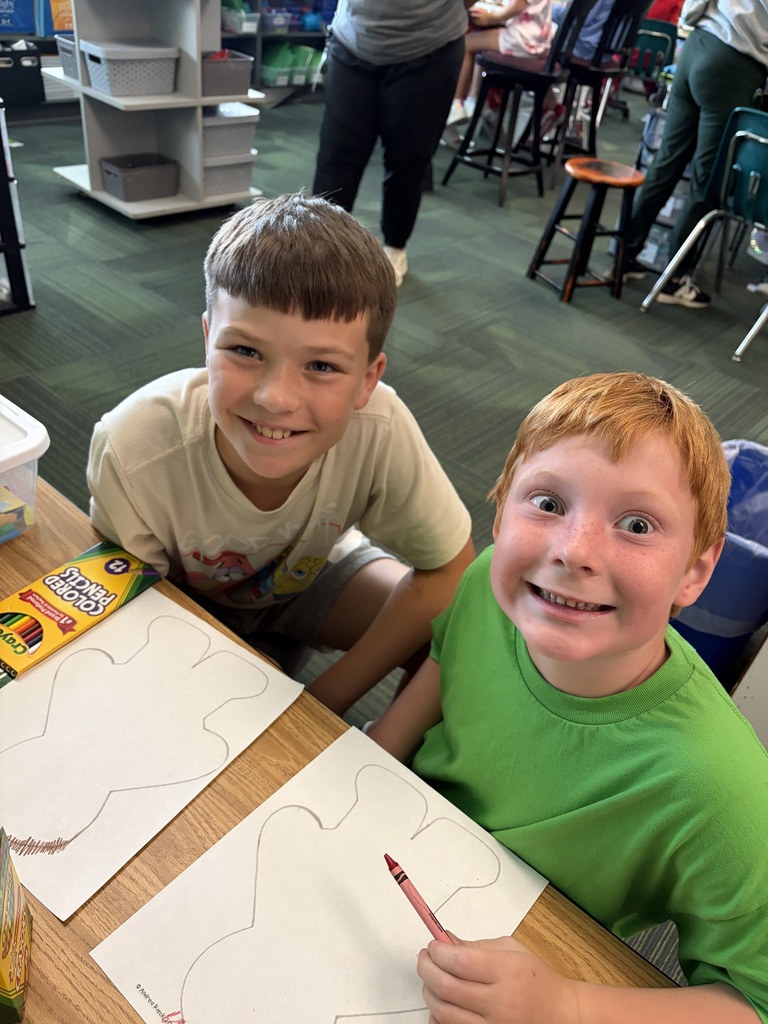 Two boys making fun faces at the camera while coloring their paper cutouts with crayons at their workspace.