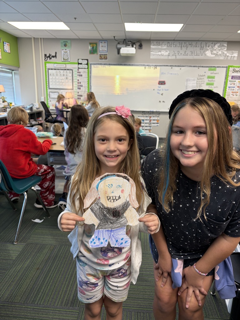 A young student proudly holding up her completed, fully colored self-portrait paper cutout alongside an older student mentor.